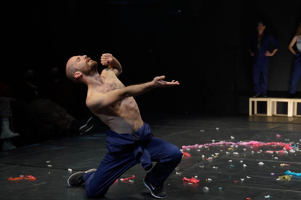 Tal kneeling on a dark stage scattered with confetti, arms reaching