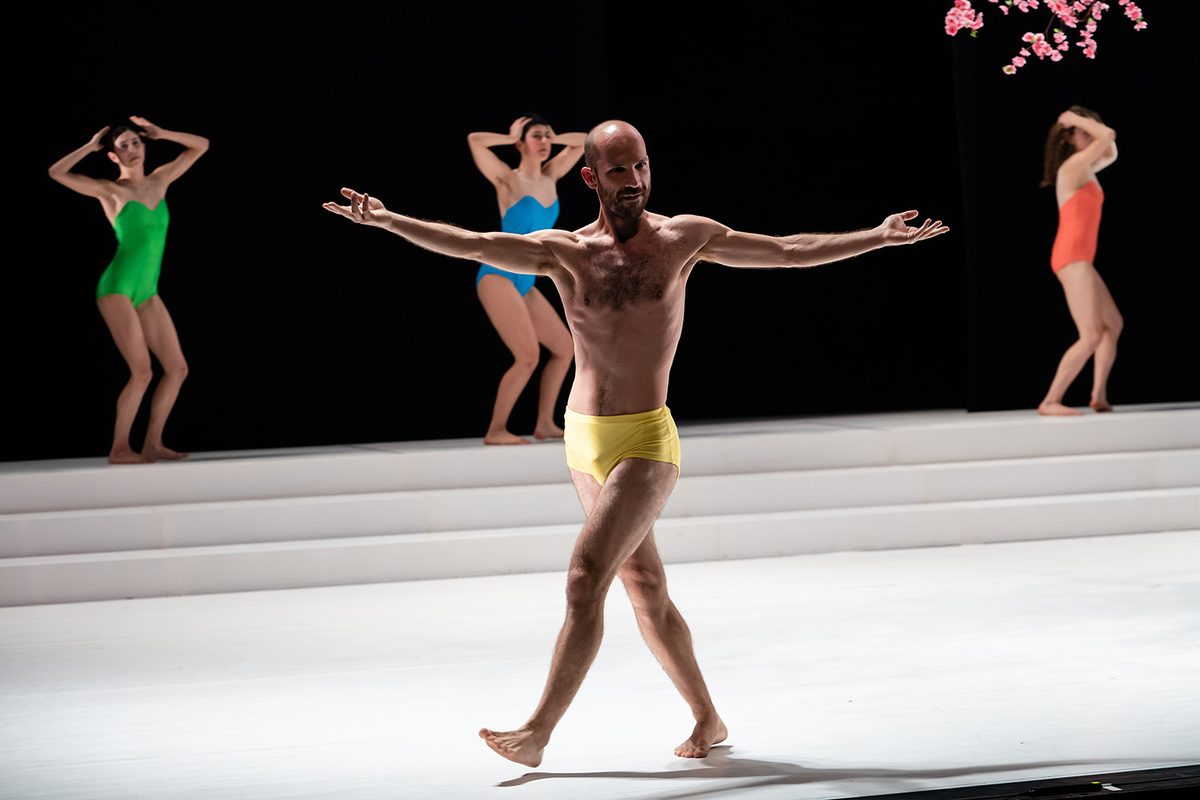 Tal walking forward with open arms on a white stage, three dancers in bright leotards behind him
